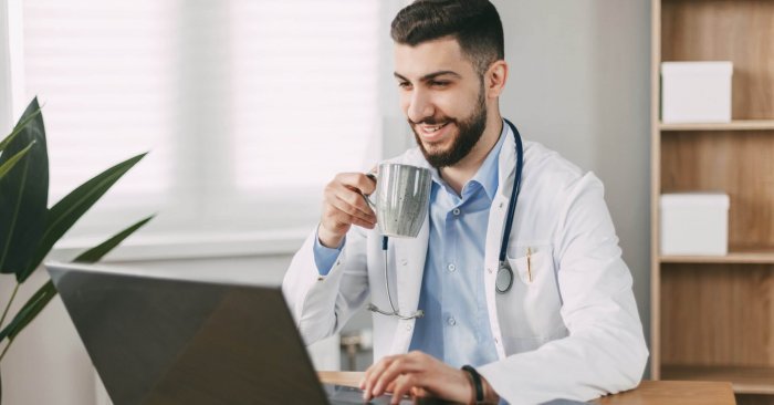 Young male doctor drinking coffee during break and communicating using laptop in his office.